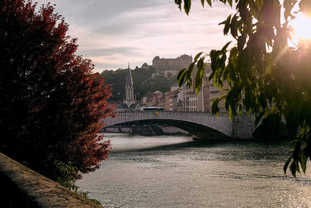 La Saône à Lyon en fin d'après-midi : un pont de pierre, les façades colorées des quais et la colline en arrière-plan, encadrés de feuillages.